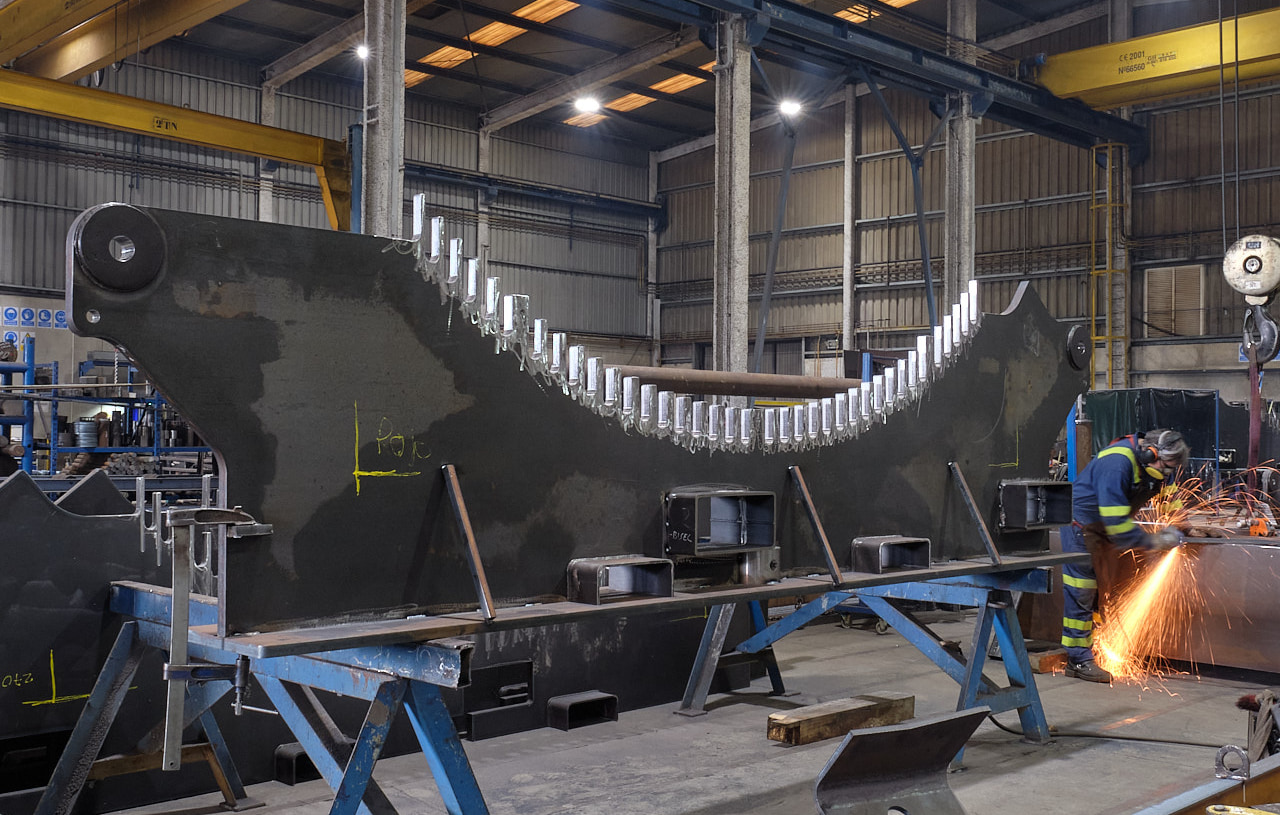 Gran pieza estructural de acero sobre caballetes azules en el taller de TMG by EPER Group, con un operario soldando al fondo