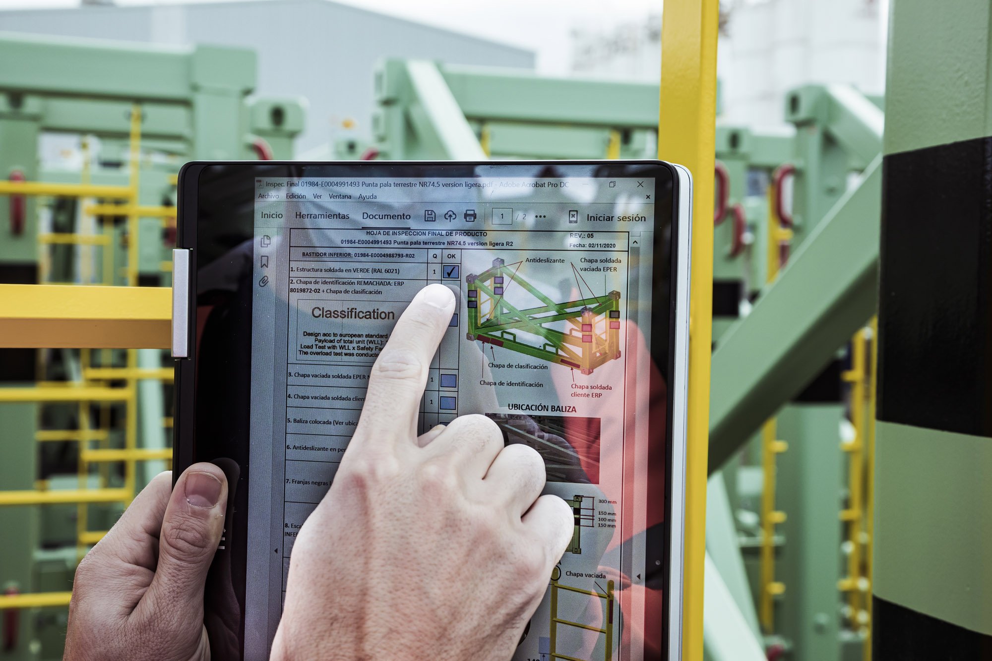 Mano sujetando una tablet con documentación de proyecto frente a una estructura metálica en el taller